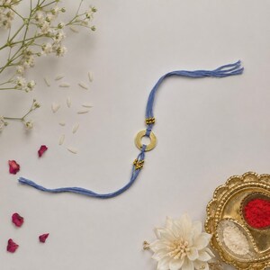 May include: A blue thread bracelet featuring a central gold-toned metal ring flanked by small gold-toned beads on each side. The bracelet is constructed from multiple strands of blue thread that extend outward from the central metal component.