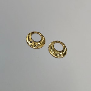 May include: A pair of gold-coloured, oval earrings. Each earring has a detailed, etched pattern of radiating lines and leaf-like shapes. The earrings have a hinged closure and are displayed on a light grey surface.