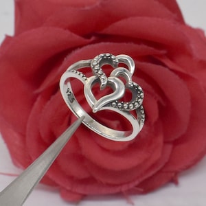 Può includere: Un anello d'argento con un design a doppio cuore. L'anello è contrassegnato dal numero "925" sulla fascia interna.