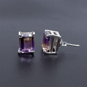 May include: A pair of stud earrings featuring rectangular, faceted amethyst gemstones. The purple and amber-toned stones are set in a silver metal frame. The earrings are displayed on a dark surface, showcasing their color and design.