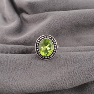 May include: A silver ring featuring a large, oval-cut, light green gemstone. The gemstone is encircled by a beaded border. The ring is set against a grey background.