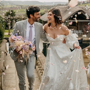 May include: A couple poses for a wedding photo, the bride in a white gown with a sweetheart neckline and the groom in a gray suit with a light purple tie. They are surrounded by falling flower petals.