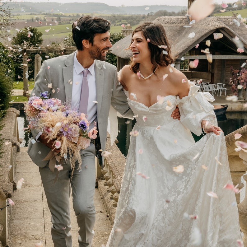 May include: A couple poses for a wedding photo, the bride in a white gown with a sweetheart neckline and the groom in a gray suit with a light purple tie. They are surrounded by falling flower petals.