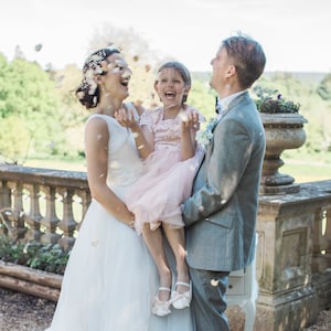 May include: A bride and groom are smiling and laughing as they hold a young girl in a pink dress. The girl is smiling and throwing flower petals in the air. The couple is standing in front of a stone wall with a flower pot on top.