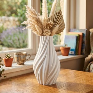 Puede incluir: Un jarrón blanco con una textura acanalada en espiral contiene un arreglo floral seco con hierba de la pampa beige, colas de conejo esponjosas y una hoja de palma en forma de abanico. El jarrón tiene un cuello estrecho que se ensancha hacia la base.