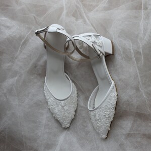 May include: A pair of white bridal shoes with delicate lace detailing and ankle straps. The shoes have a low block heel and a pointed toe.