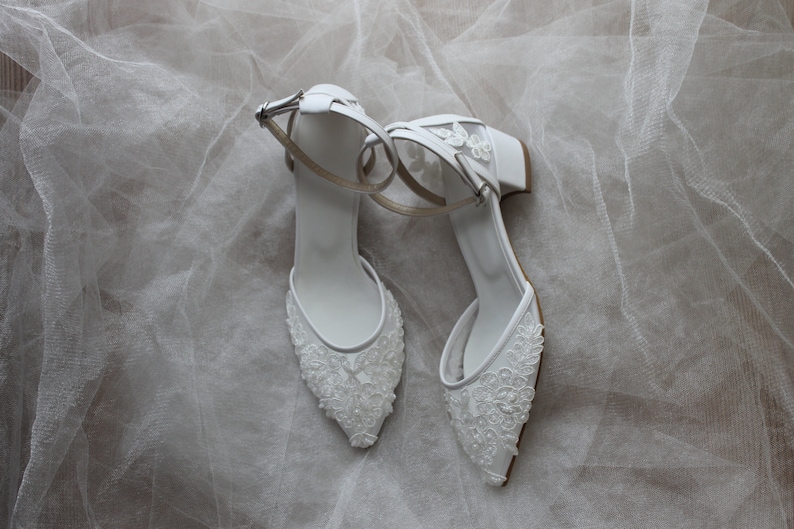 May include: A pair of white bridal shoes with delicate lace detailing and ankle straps. The shoes have a low block heel and a pointed toe.