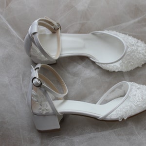 May include: A pair of white bridal shoes with delicate lace detailing and ankle straps. The shoes have a low heel and a pointed toe.