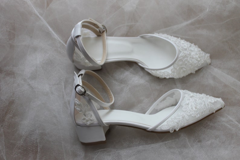 May include: A pair of white bridal shoes with delicate lace detailing and ankle straps. The shoes have a low heel and a pointed toe.