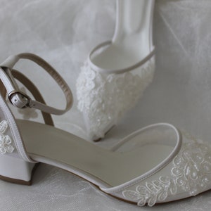 May include: White satin bridal shoes with delicate lace and pearl embellishments. The shoes have a low block heel and an ankle strap with a buckle.