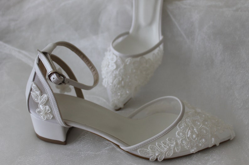 May include: White satin bridal shoes with delicate lace and pearl embellishments. The shoes have a low block heel and an ankle strap with a buckle.
