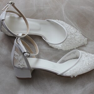May include: A pair of white bridal shoes with delicate lace detailing. The shoes have a low heel and an ankle strap.