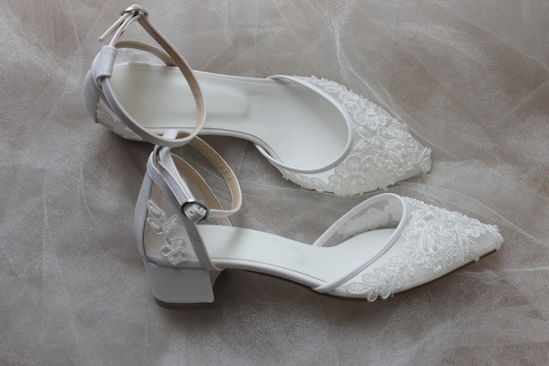 May include: A pair of white bridal shoes with delicate lace detailing. The shoes have a low heel and an ankle strap.