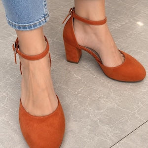 May include: A pair of burnt orange suede-textured block heel shoes with a rounded toe and an adjustable ankle strap featuring a silver-toned metal buckle. The shoes have a closed heel design and a sturdy, medium-height block heel for support.