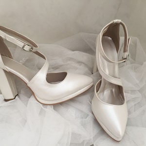 Pu&ograve; includere: Un paio di scarpe da sposa in raso bianco con punta a punta e tacco a blocco spesso. Le scarpe hanno un delicato cinturino alla caviglia con chiusura a fibbia.