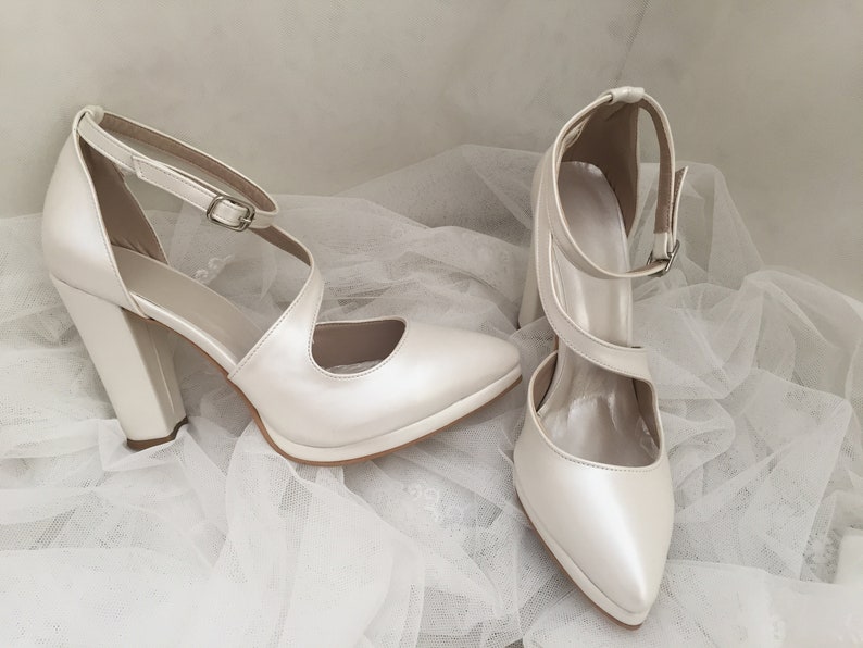 Pu&ograve; includere: Un paio di scarpe da sposa in raso bianco con punta a punta e tacco a blocco spesso. Le scarpe hanno un delicato cinturino alla caviglia con chiusura a fibbia.