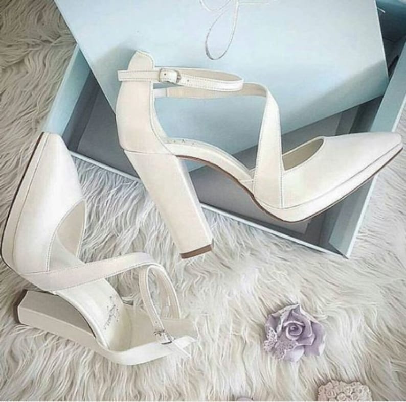 Pu&ograve; includere: Un paio di scarpe da sposa in raso bianco con un tacco spesso e un cinturino alla caviglia. Le scarpe sono in una scatola con una fodera blu chiaro.