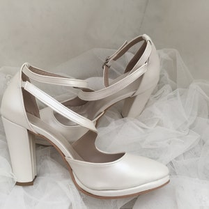 Pu&ograve; includere: Un paio di tacchi da sposa in raso bianco con un tacco a blocco spesso e un cinturino alla caviglia. I tacchi sono su un tessuto di tulle bianco.