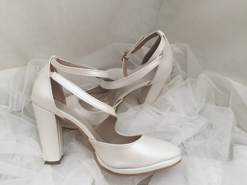 Pu&ograve; includere: Un paio di tacchi da sposa in raso bianco con un tacco a blocco spesso e un cinturino alla caviglia. I tacchi sono su un tessuto di tulle bianco.