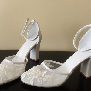 Pode incluir: Um par de sapatos de noiva brancos com uma delicada sobreposi&ccedil;&atilde;o de renda e um salto bloco.