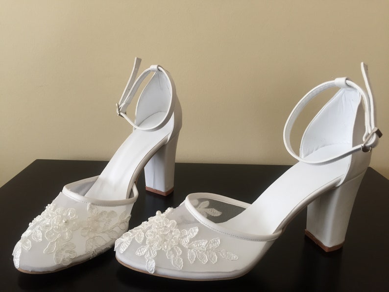 Pode incluir: Um par de sapatos de noiva brancos com uma delicada sobreposi&ccedil;&atilde;o de renda e um salto bloco.