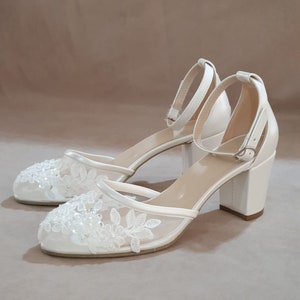 Pode incluir: Sapatos de noiva de cetim branco com uma delicada sobreposi&ccedil;&atilde;o de renda e um salto bloco. Os sapatos t&ecirc;m uma tira de tornozelo com fecho de fivela.