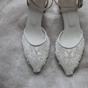 May include: A pair of white bridal shoes with delicate lace detailing and a pointed toe.