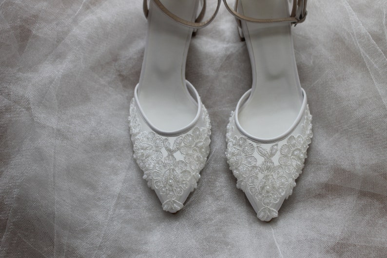 May include: A pair of white bridal shoes with delicate lace detailing and a pointed toe.