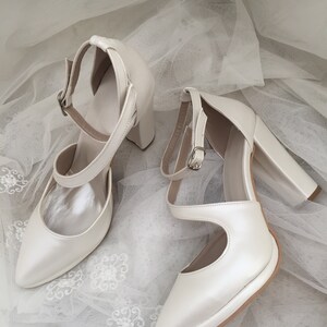 Pu&ograve; includere: Un paio di scarpe da sposa in raso bianco con punta a punta e tacco spesso. Le scarpe hanno un delicato cinturino alla caviglia con fibbia.