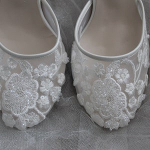 Pode incluir: Um par de sapatos de noiva brancos com detalhes delicados em renda. Os sapatos t&ecirc;m bico fino e salto baixo.