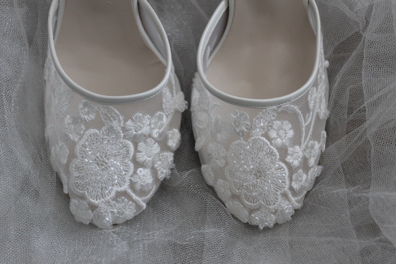 Pode incluir: Um par de sapatos de noiva brancos com detalhes delicados em renda. Os sapatos t&ecirc;m bico fino e salto baixo.