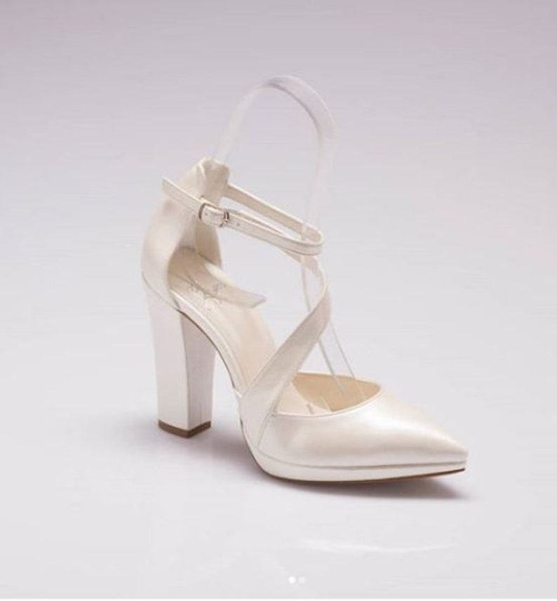 Pu&ograve; includere: Scarpe da sposa in raso bianco con punta a punta e tacco a blocco spesso. Le scarpe hanno un design a cinturino incrociato e una chiusura a fibbia.