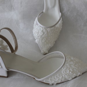 May include: A pair of white bridal shoes with delicate floral lace detailing. The shoes have a low block heel and an ankle strap.