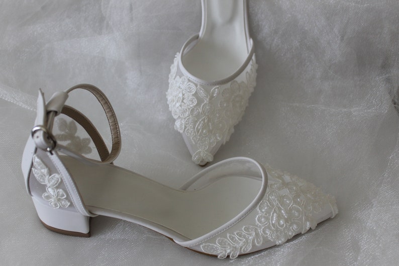 May include: A pair of white bridal shoes with delicate floral lace detailing. The shoes have a low block heel and an ankle strap.
