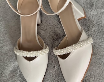 May include: A pair of white pointed-toe heels with ankle straps and a small block heel. The straps are secured with a buckle. The upper part of the shoe is decorated with a row of small pearls.
