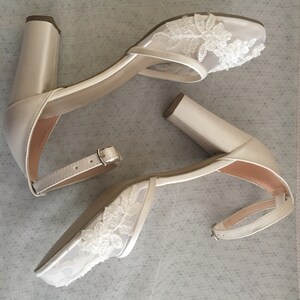 Pode incluir: Um par de sapatos de noiva de cetim branco com detalhes delicados de renda nos dedos. Os saltos t&ecirc;m um salto de bloco e uma tira de tornozelo.