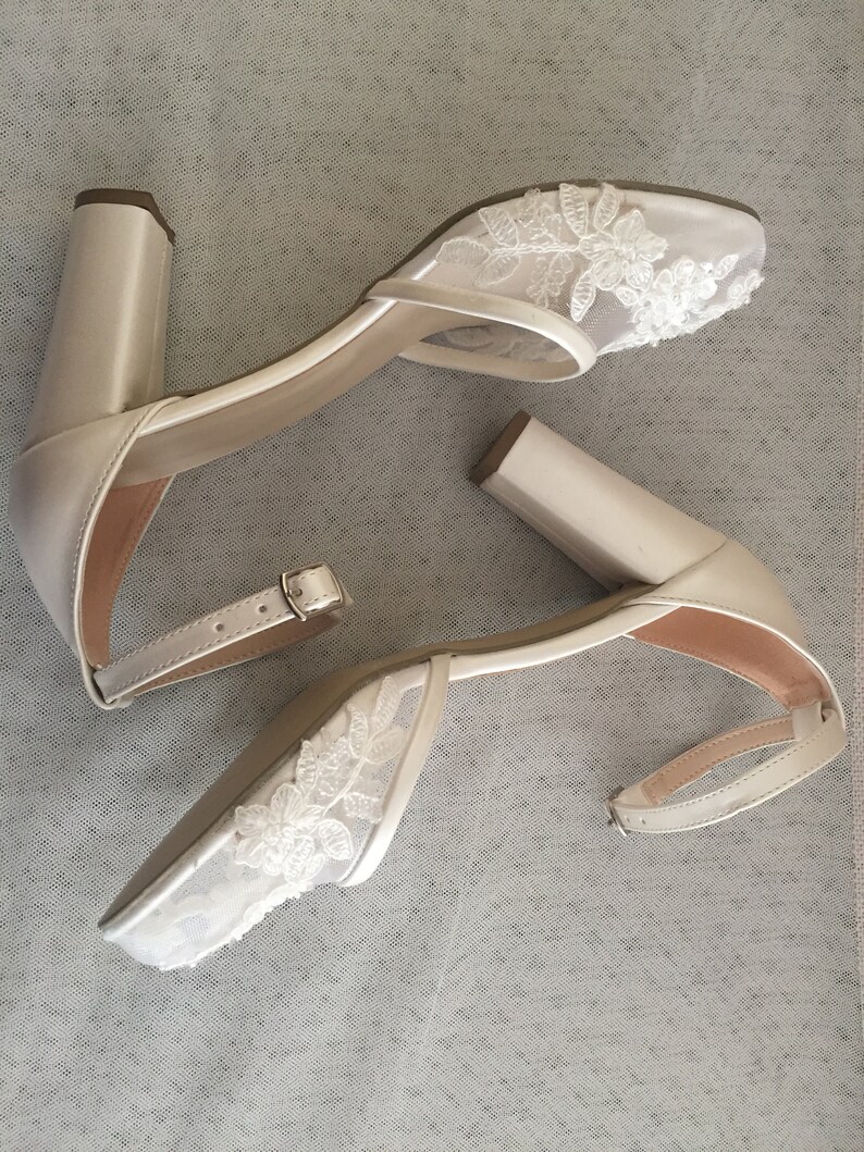Pode incluir: Um par de sapatos de noiva de cetim branco com detalhes delicados de renda nos dedos. Os saltos t&ecirc;m um salto de bloco e uma tira de tornozelo.