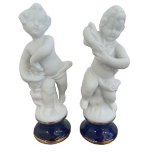 Könnte beinhalten: Zwei weiße Porzellan-Engelfiguren mit blauen und goldenen Sockeln. Die Engel sind spielerisch posiert, einer hält einen Blumenkorb und der andere ein Musikinstrument.