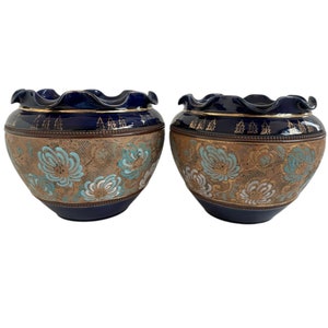 May include: A pair of blue ceramic bowls with a decorative floral design. The bowls have a ruffled rim with gold detailing. The body of the bowls features a pattern of blue, gold, and white flowers. The bowls are set against a white background.