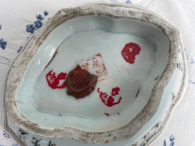 May include: Antique porcelain dish with a light blue interior and a textured, irregular rim. The dish features red markings and a brown wax seal. The bottom of the dish has a light blue color with some text.