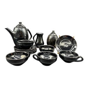 Peut inclure: Un service à thé complet en céramique noire, comprenant une théière, des tasses, des soucoupes, des bols, une crémière, un sucrier et une assiette de service. L'ensemble a une finition brillante et est disposé sur fond blanc. Le service à thé est de conception classique.