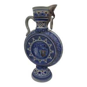 Può includere: Una brocca in ceramica circolare e alta con base grigio chiaro e motivi in rilievo blu cobalto. Il medaglione centrale raffigura una figura e uno scudo, mentre il collo presenta un manico decorativo e un elemento ad anello.