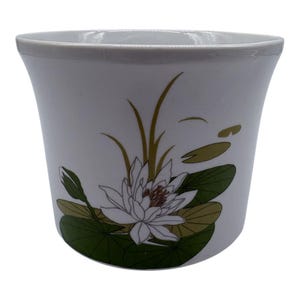 Peut inclure: Pot de fleurs en céramique blanche avec une forme évasée et un bord gris. Le pot présente un motif floral avec un nénuphar blanc, des feuilles de nénuphar vertes et de longues tiges fines vertes et dorées.