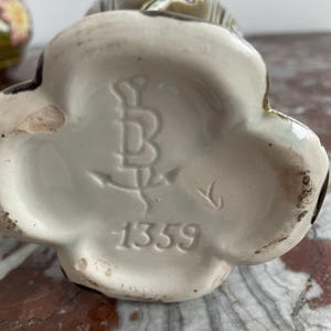 May include: Close-up of the bottom of a ceramic vase with a floral design. The vase has a white glaze and a brown mark with the letter "B" and the number "1359".