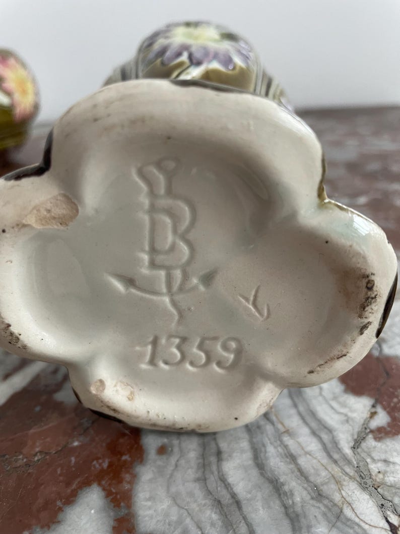 May include: Close-up of the bottom of a ceramic vase with a floral design. The vase has a white glaze and a brown mark with the letter "B" and the number "1359".