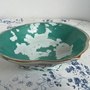 May include: A decorative, antique ceramic bowl with a scalloped edge. The bowl features a teal interior with white accents and a brown rim. The base has a floral design. The bowl is likely used for serving or display.