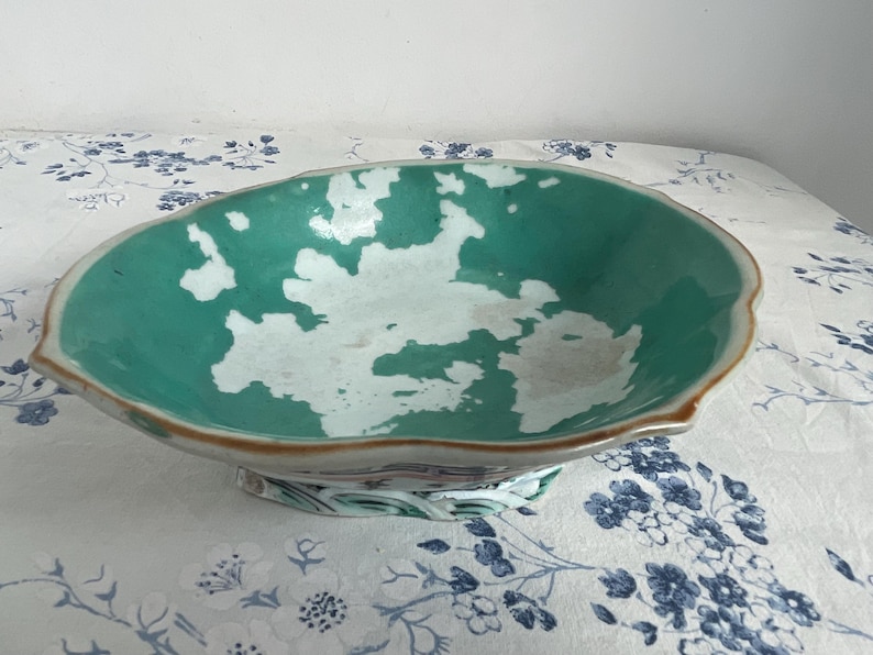 May include: A decorative, antique ceramic bowl with a scalloped edge. The bowl features a teal interior with white accents and a brown rim. The base has a floral design. The bowl is likely used for serving or display.