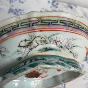 May include: A detailed view of a porcelain bowl with a scalloped rim. The bowl features hand-painted floral and landscape designs in shades of green, pink, and white. The rim has a decorative border with geometric patterns. The bowl is set against a light blue background.