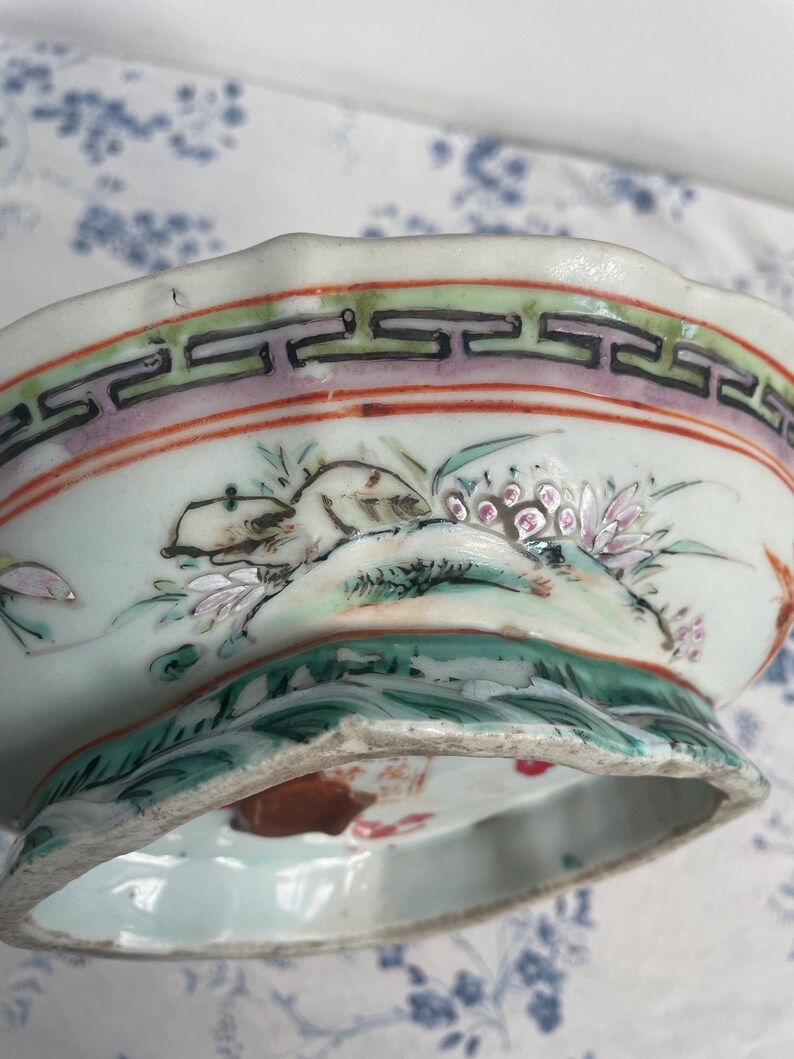 May include: A detailed view of a porcelain bowl with a scalloped rim. The bowl features hand-painted floral and landscape designs in shades of green, pink, and white. The rim has a decorative border with geometric patterns. The bowl is set against a light blue background.