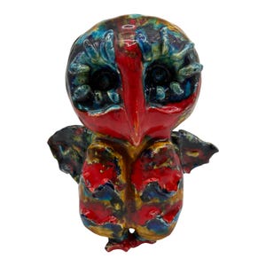 May include: A ceramic owl figurine featuring a glossy, multi-colored glaze in vibrant shades of red, deep blue, and golden yellow. The owl has large, dark, recessed eyes surrounded by textured, raised blue ridges and a prominent red beak.